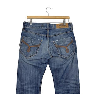 K&ouml;nnte beinhalten: Blaue Jeans aus Denim mit Used-Details und dekorativen N&auml;hten auf den Ges&auml;&szlig;taschen. Die Jeans h&auml;ngen an einem Holzb&uuml;gel. Ein Lederetikett ist auf der R&uuml;ckseite der Jeans sichtbar.