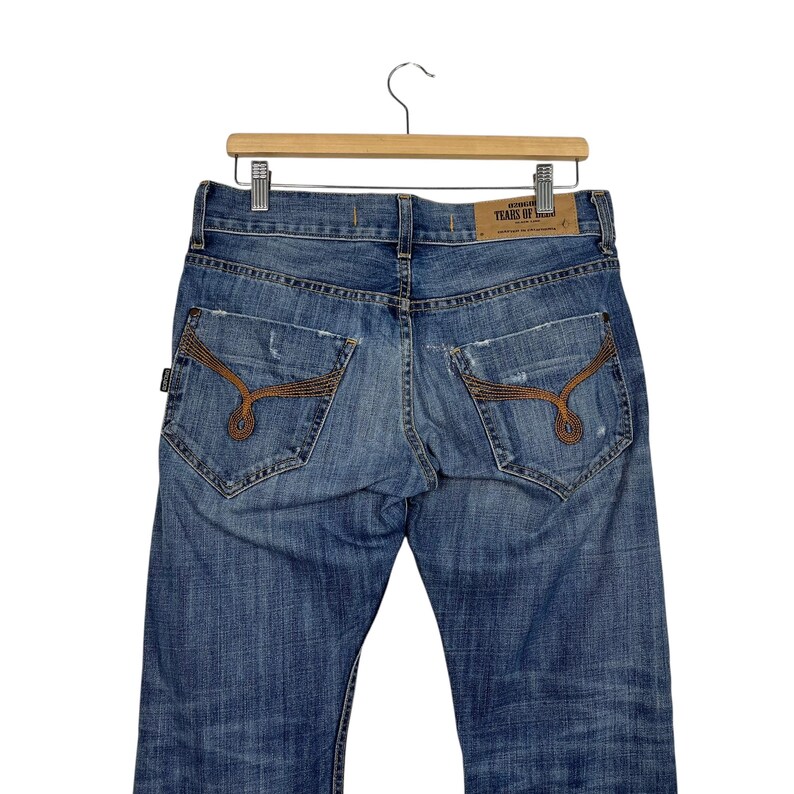 K&ouml;nnte beinhalten: Blaue Jeans aus Denim mit Used-Details und dekorativen N&auml;hten auf den Ges&auml;&szlig;taschen. Die Jeans h&auml;ngen an einem Holzb&uuml;gel. Ein Lederetikett ist auf der R&uuml;ckseite der Jeans sichtbar.