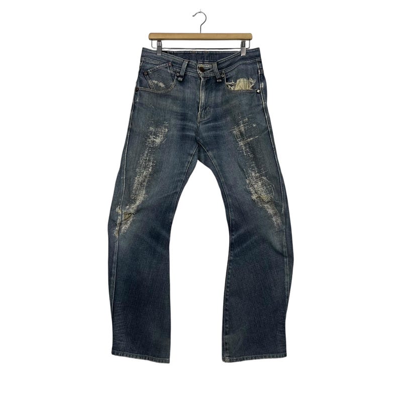 K&ouml;nnte beinhalten: Dunkelblaue Jeans mit Used-Details, h&auml;ngen an einem Holzkleiderb&uuml;gel. Die Jeans haben ein klassisches F&uuml;nf-Taschen-Design und ein leicht ausgestelltes Bein. Die Used-Bereiche haben ein verwaschenes Aussehen.