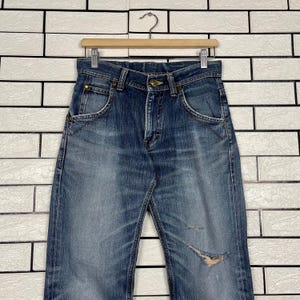 May include: Blue denim jeans with a faded wash and distressed details, hanging on a wooden hanger. The jeans have classic five-pocket styling and a button and zipper closure. The jeans are approximately 32 inches long.