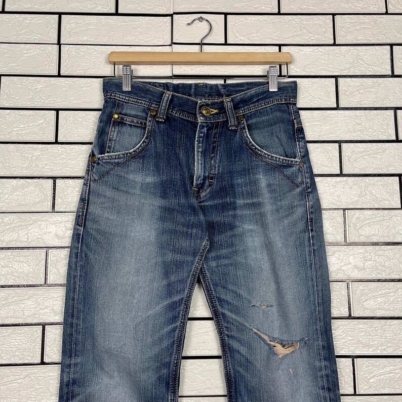 May include: Blue denim jeans with a faded wash and distressed details, hanging on a wooden hanger. The jeans have classic five-pocket styling and a button and zipper closure. The jeans are approximately 32 inches long.