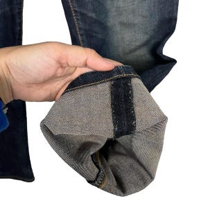 Puede incluir: Primer plano de unos vaqueros de mezclilla de lavado oscuro. Los vaqueros est&aacute;n doblados, revelando la tela interior y las costuras de color m&aacute;s claro. La mezclilla tiene un aspecto desgastado.