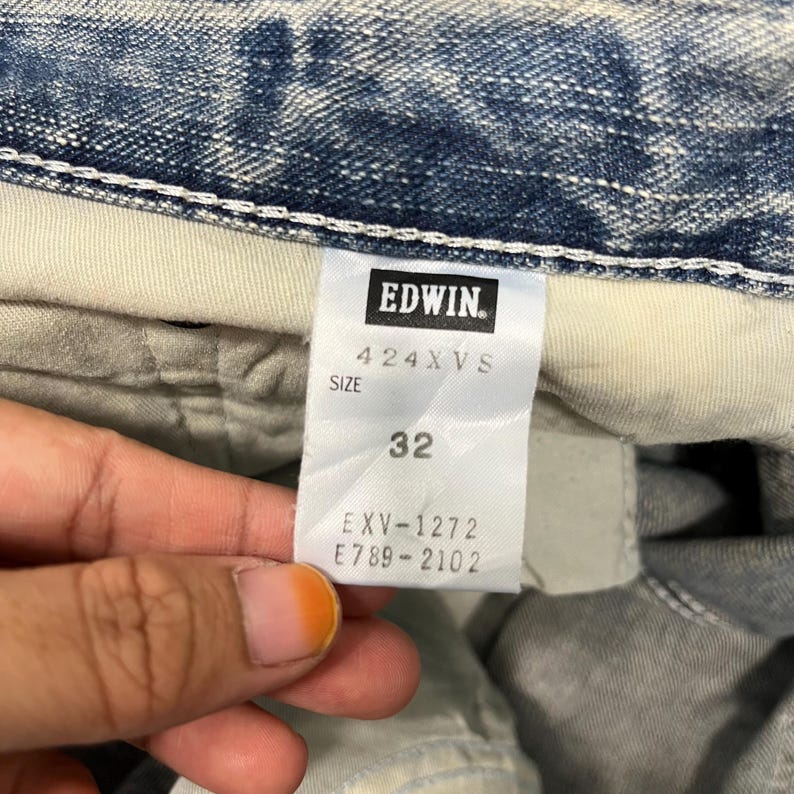 May include: Close-up of a pair of blue denim jeans with a white tag. The tag features the brand name "Edwin" in a black rectangle, along with the size "32" and other product information.