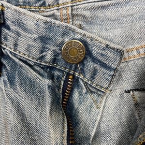 May include: Close-up of light blue denim jeans with a brass button and zipper. The button is embossed with the words "EDWIN CO LTD". The jeans have visible stitching and a classic design.