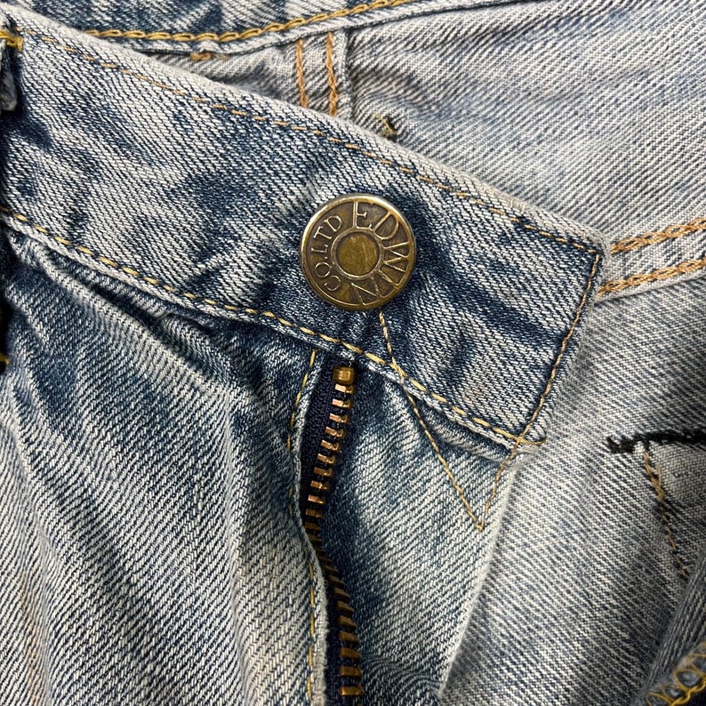 May include: Close-up of light blue denim jeans with a brass button and zipper. The button is embossed with the words "EDWIN CO LTD". The jeans have visible stitching and a classic design.