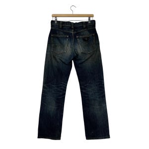 May include: Dark wash denim jeans hanging on a wooden hanger. The jeans have a classic five-pocket design, a button and zipper closure, and a faded, worn appearance. The back pockets feature a decorative design.
