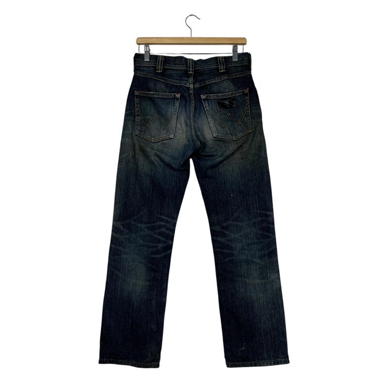 May include: Dark wash denim jeans hanging on a wooden hanger. The jeans have a classic five-pocket design, a button and zipper closure, and a faded, worn appearance. The back pockets feature a decorative design.