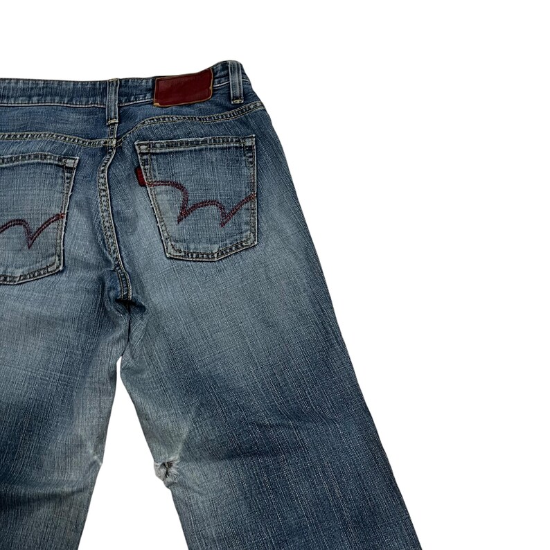 Op de afbeelding: Verbleekte blauwe spijkerbroek met rode stiksels op de achterzakken en een bruine leren patch. De jeans heeft een klassiek ontwerp met vijf zakken en een relaxte pasvorm. De denim heeft een versleten look.