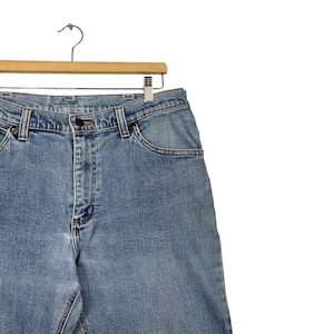 K&ouml;nnte beinhalten: Hellblaue Jeans aus Denim, die an einem Holzkleiderb&uuml;gel h&auml;ngen. Die Jeans haben ein klassisches F&uuml;nf-Taschen-Design mit Knopf- und Rei&szlig;verschluss. Der Denim hat ein verwaschenes Aussehen.