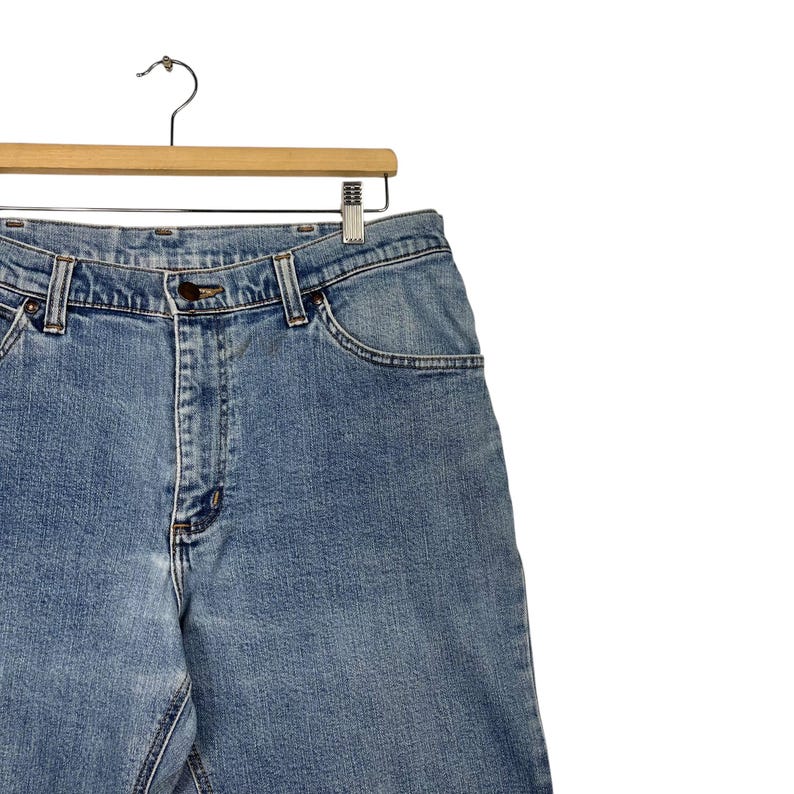 K&ouml;nnte beinhalten: Hellblaue Jeans aus Denim, die an einem Holzkleiderb&uuml;gel h&auml;ngen. Die Jeans haben ein klassisches F&uuml;nf-Taschen-Design mit Knopf- und Rei&szlig;verschluss. Der Denim hat ein verwaschenes Aussehen.
