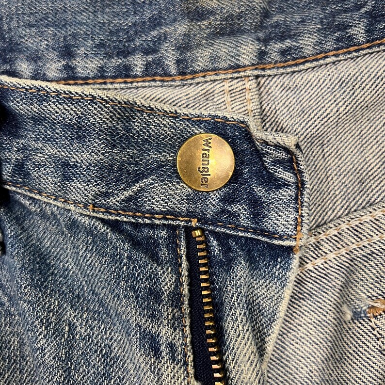 K&ouml;nnte beinhalten: Nahaufnahme einer blauen Jeans mit einem messingfarbenen Knopf mit der Aufschrift "Wrangler". Der Rei&szlig;verschluss ist sichtbar, und der Denim hat ein abgenutztes, verwaschenes Aussehen. Die N&auml;hte sind hellbraun.