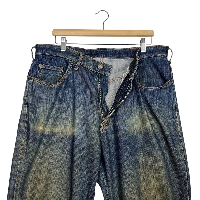K&ouml;nnte beinhalten: Blaue Jeans mit verwaschener Optik, h&auml;ngen an einem Holzb&uuml;gel. Die Jeans haben einen Knopf- und Rei&szlig;verschluss, Vordertaschen und ein klassisches F&uuml;nf-Taschen-Design. Der Denim weist Gebrauchsspuren auf.