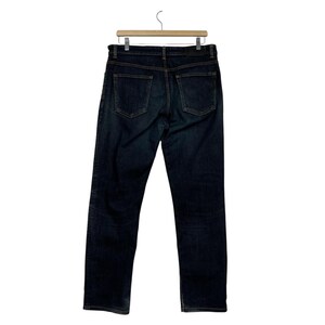 Puede incluir: Vaqueros de mezclilla de color azul oscuro colgados de una percha de madera. Los vaqueros tienen un dise&ntilde;o cl&aacute;sico de cinco bolsillos con corte recto. Los bolsillos traseros son visibles y las costuras son de color contrastante.