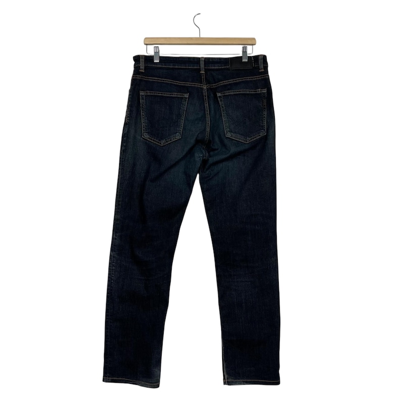 Puede incluir: Vaqueros de mezclilla de color azul oscuro colgados de una percha de madera. Los vaqueros tienen un dise&ntilde;o cl&aacute;sico de cinco bolsillos con corte recto. Los bolsillos traseros son visibles y las costuras son de color contrastante.