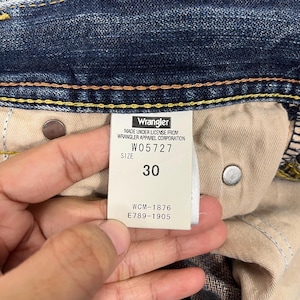 May include: Close-up of a pair of blue denim Wrangler jeans. The tag reads "Wrangler" and includes the size "30". The tag also displays the style number W05727 and other manufacturing codes.