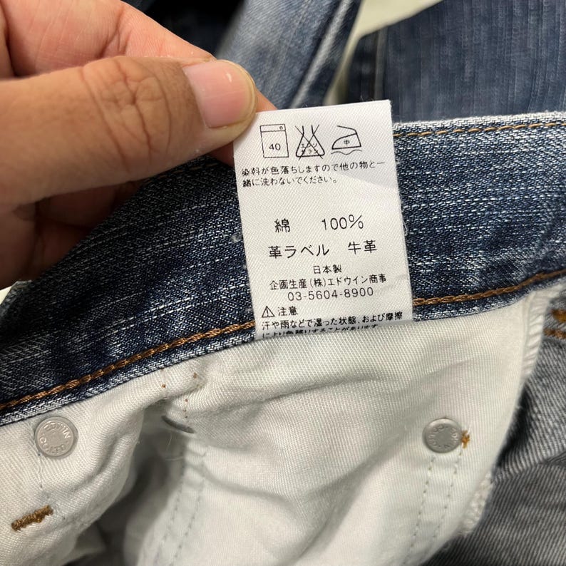 May include: A close-up of a denim jeans label with Japanese text. The label shows washing instructions, fabric composition (100% cotton), and manufacturer details. The jeans are a medium wash blue.