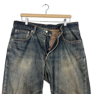 May include: Blue denim jeans with a faded wash, hanging on a wooden hanger. The jeans have a button and zipper closure, with visible pocket details. The fabric shows signs of wear and tear.