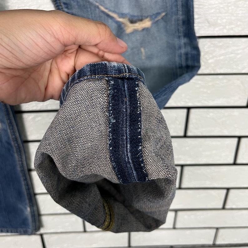 May include: Close-up of the hem of a pair of blue denim jeans. The jeans have a distressed look with a faded wash and a torn section. The hem is folded up, revealing the inside fabric.