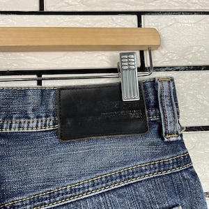 May include: Close-up of blue denim jeans with a black leather patch and yellow stitching. The jeans are hanging on a wooden hanger with a silver metal clip. The denim has a faded appearance.