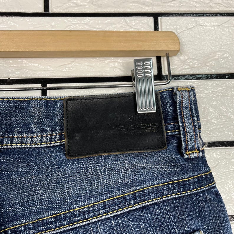 May include: Close-up of blue denim jeans with a black leather patch and yellow stitching. The jeans are hanging on a wooden hanger with a silver metal clip. The denim has a faded appearance.