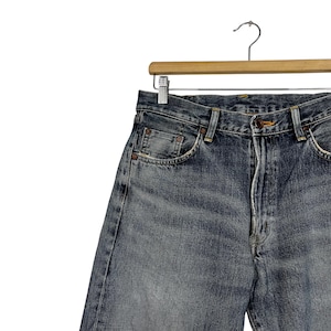 May include: A pair of blue denim jeans with a faded wash. The jeans are hanging on a wooden hanger.