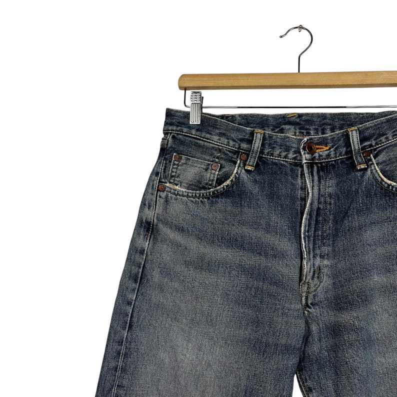 May include: A pair of blue denim jeans with a faded wash. The jeans are hanging on a wooden hanger.