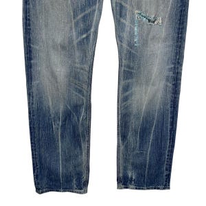May include: Faded blue denim jeans with distressed detailing. The jeans have a worn appearance with visible fading and a ripped section on the right leg, patched with a lighter blue fabric. The jeans are a classic five-pocket style.