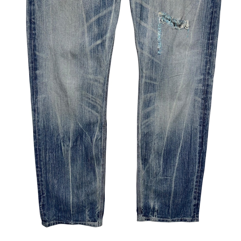 May include: Faded blue denim jeans with distressed detailing. The jeans have a worn appearance with visible fading and a ripped section on the right leg, patched with a lighter blue fabric. The jeans are a classic five-pocket style.