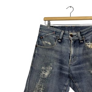K&ouml;nnte beinhalten: Blaue Jeans aus Denim mit Used-Details, h&auml;ngen an einem Holzb&uuml;gel. Die Jeans haben einen Knopfverschluss, Vordertaschen und sichtbare Abnutzungserscheinungen. Der Denim hat ein verwaschenes Aussehen.