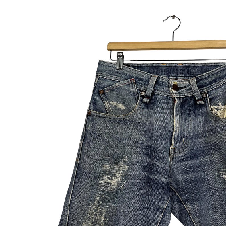 K&ouml;nnte beinhalten: Blaue Jeans aus Denim mit Used-Details, h&auml;ngen an einem Holzb&uuml;gel. Die Jeans haben einen Knopfverschluss, Vordertaschen und sichtbare Abnutzungserscheinungen. Der Denim hat ein verwaschenes Aussehen.