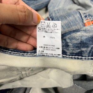May include: A close-up of a tag with Japanese text and care symbols. The tag reads "綿 100%" and "MADE IN CHINA". The tag is attached to a pair of blue denim jeans.