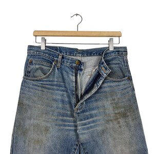 K&ouml;nnte beinhalten: Hellblaue Jeans mit verblichener Waschung und sichtbaren Gebrauchsspuren, h&auml;ngen an einem Holzkleiderb&uuml;gel. Die Jeans sind aufgekn&ouml;pft und ge&ouml;ffnet. Sie haben Vordertaschen und ein klassisches F&uuml;nf-Taschen-Design.