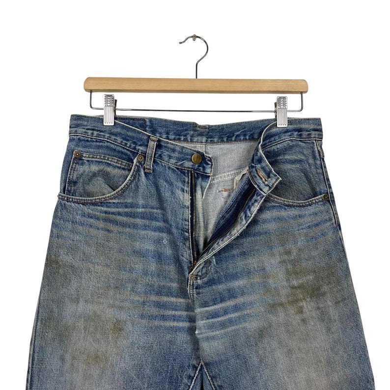 K&ouml;nnte beinhalten: Hellblaue Jeans mit verblichener Waschung und sichtbaren Gebrauchsspuren, h&auml;ngen an einem Holzkleiderb&uuml;gel. Die Jeans sind aufgekn&ouml;pft und ge&ouml;ffnet. Sie haben Vordertaschen und ein klassisches F&uuml;nf-Taschen-Design.