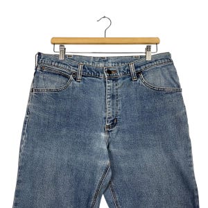 K&ouml;nnte beinhalten: Hellblaue Jeans an einem Holzb&uuml;gel. Die Jeans haben ein klassisches F&uuml;nf-Taschen-Design mit Knopf- und Rei&szlig;verschluss. Der Denim hat ein verwaschenes Aussehen, was auf Abnutzung und Waschen hindeutet.