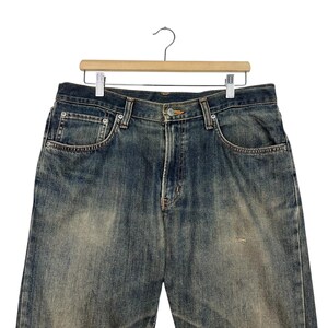May include: Faded blue denim jeans hanging on a wooden hanger. The jeans have a classic five-pocket design with a button and zipper closure. The denim has a worn, distressed look with faded areas and subtle wear marks.
