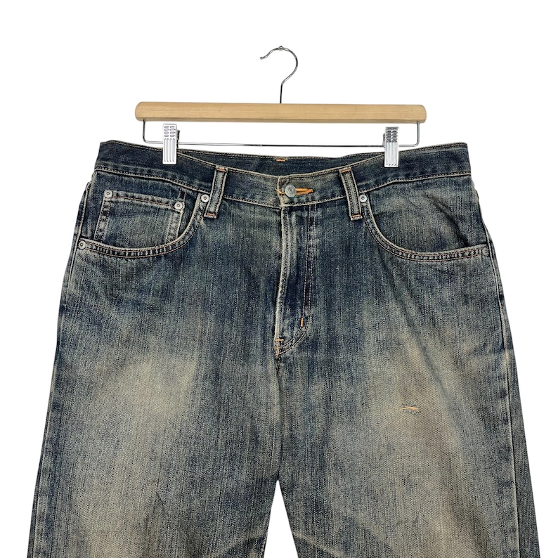 May include: Faded blue denim jeans hanging on a wooden hanger. The jeans have a classic five-pocket design with a button and zipper closure. The denim has a worn, distressed look with faded areas and subtle wear marks.