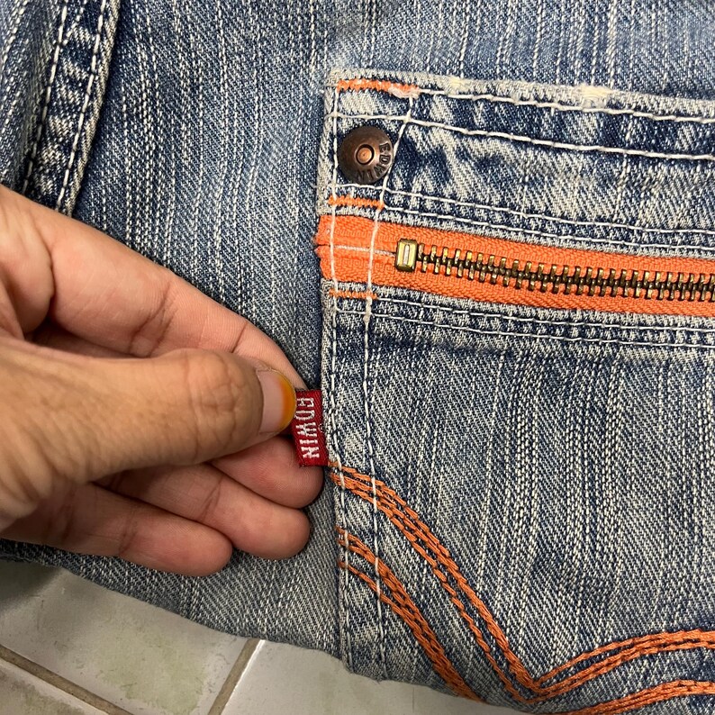 May include: Close-up of denim jeans with an orange zipper pocket and stitching. The jeans have a faded blue wash and a small red Edwin tag.
