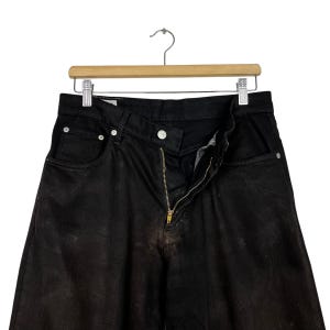 May include: Black denim jeans with a brass zipper and button closure, hanging on a wooden hanger. The jeans have a faded appearance and classic five-pocket design. The jeans are a dark wash.