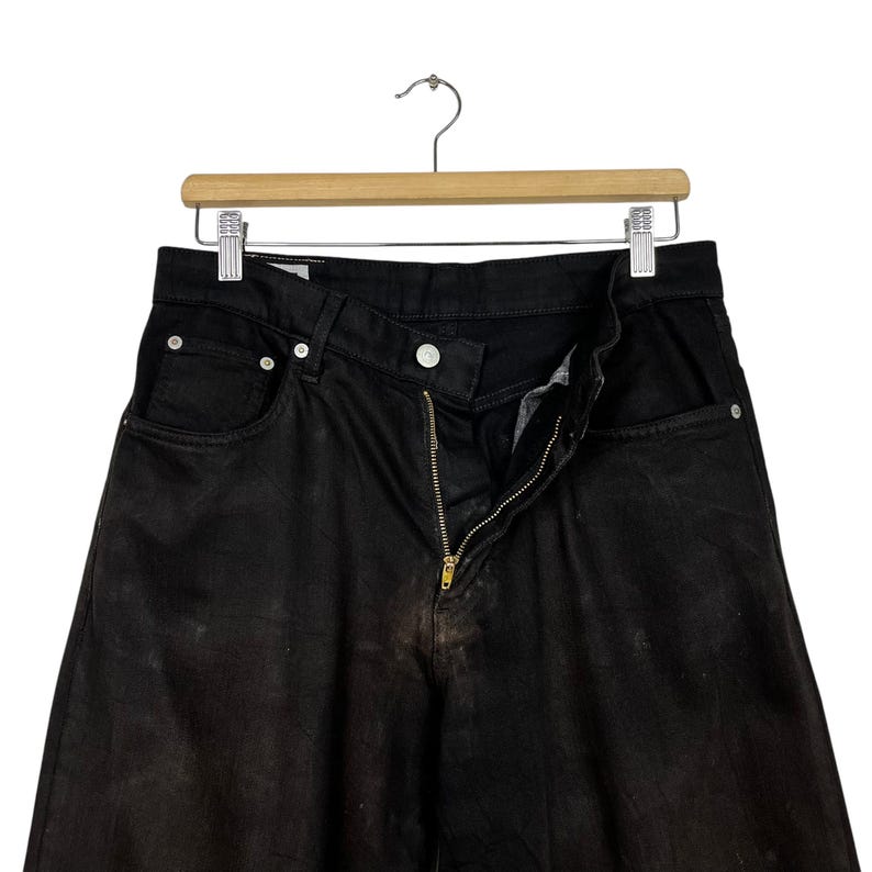 May include: Black denim jeans with a brass zipper and button closure, hanging on a wooden hanger. The jeans have a faded appearance and classic five-pocket design. The jeans are a dark wash.