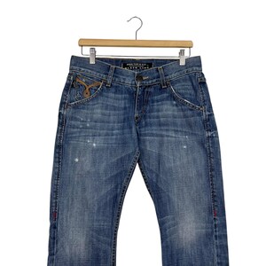 K&ouml;nnte beinhalten: Blaue Jeans aus Denim mit verwaschener Optik, mit klassischem F&uuml;nf-Taschen-Design. Die Jeans hat einen Knopf- und Rei&szlig;verschluss, mit dekorativen N&auml;hten und einem kleinen gestickten Detail auf der Vordertasche. Die Jeans h&auml;ngt an einem Holzb&uuml;gel.