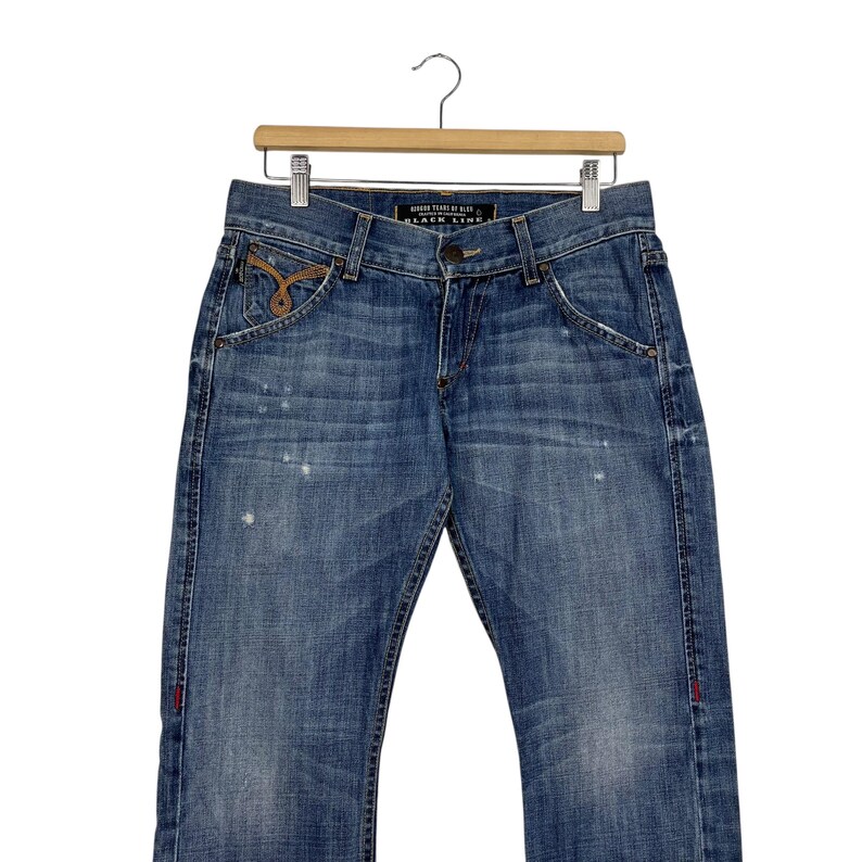 K&ouml;nnte beinhalten: Blaue Jeans aus Denim mit verwaschener Optik, mit klassischem F&uuml;nf-Taschen-Design. Die Jeans hat einen Knopf- und Rei&szlig;verschluss, mit dekorativen N&auml;hten und einem kleinen gestickten Detail auf der Vordertasche. Die Jeans h&auml;ngt an einem Holzb&uuml;gel.