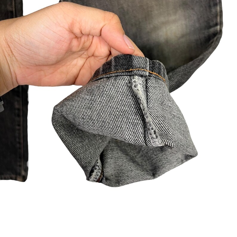 Puede incluir: Primer plano de unos vaqueros de mezclilla grises. El dobladillo est&aacute; doblado, revelando la costura interior y las costuras. La mezclilla tiene un aspecto descolorido, con un ligero degradado de color. La tela tiene un aspecto texturizado.