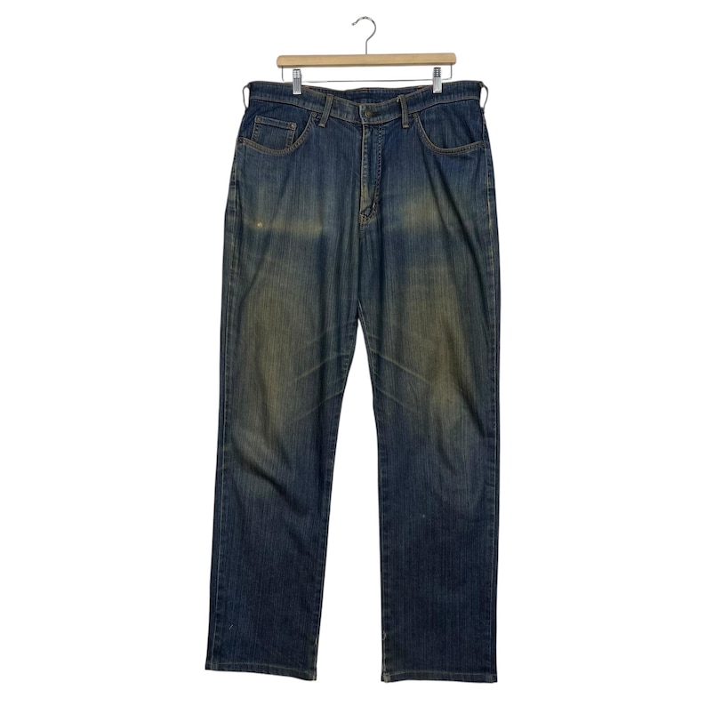 K&ouml;nnte beinhalten: Blaue Jeans aus Denim mit verwaschener Optik, h&auml;ngen an einem Holzb&uuml;gel. Die Jeans haben ein klassisches F&uuml;nf-Taschen-Design und einen geraden Beinverlauf. Der Denim hat ein abgenutztes Aussehen mit helleren Bereichen.