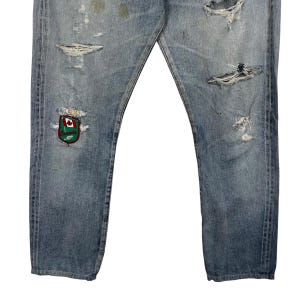 May include: Light blue denim jeans with distressed details, including rips and patches. Features a colorful embroidered crest with a maple leaf design. The jeans have a classic five-pocket design.