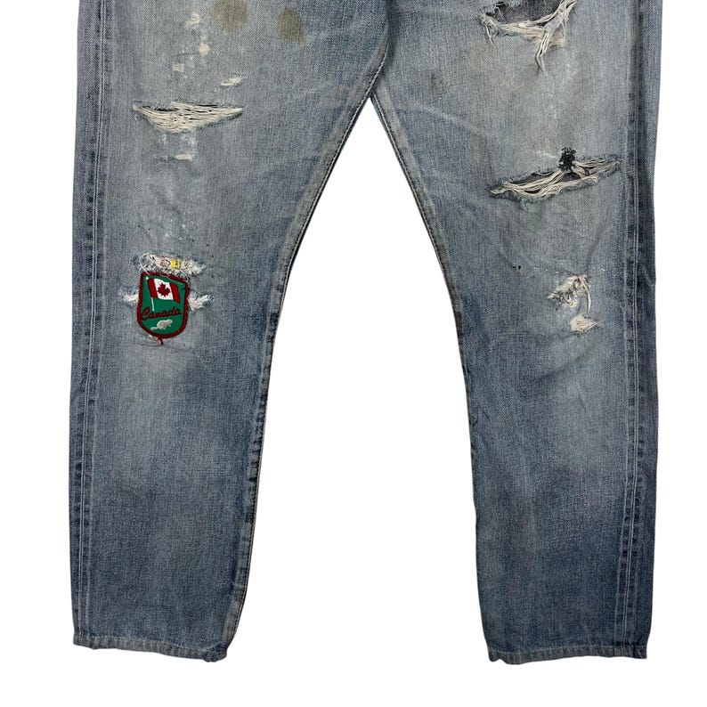 May include: Light blue denim jeans with distressed details, including rips and patches. Features a colorful embroidered crest with a maple leaf design. The jeans have a classic five-pocket design.
