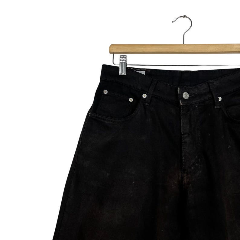 May include: Close-up of black denim jeans hanging on a wooden hanger. The jeans have a classic five-pocket design with a button and zipper closure. The fabric appears slightly faded, adding a vintage look.
