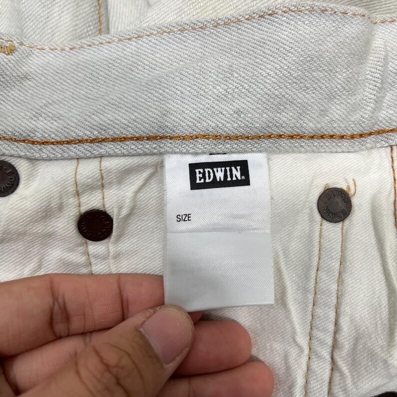 May include: Close-up of the inside of light-colored denim jeans. A white tag with the brand name "EDWIN" in black lettering is visible, along with the word "SIZE". Brown metal buttons and stitching are also present.