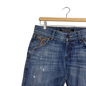 K&ouml;nnte beinhalten: Blaue Jeans aus Denim mit dekorativem Taschen-Design und goldfarbenen Stickereien. Die Jeans h&auml;ngen an einem Holzb&uuml;gel. Der Text "BLACK LINE" ist auf dem Bund zu sehen.