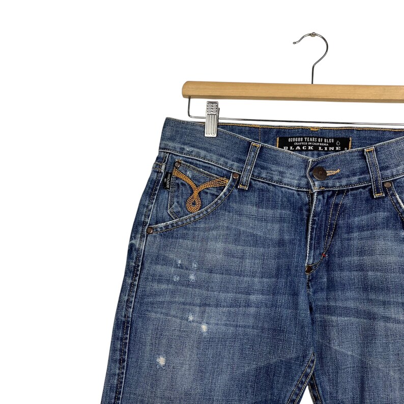 K&ouml;nnte beinhalten: Blaue Jeans aus Denim mit dekorativem Taschen-Design und goldfarbenen Stickereien. Die Jeans h&auml;ngen an einem Holzb&uuml;gel. Der Text "BLACK LINE" ist auf dem Bund zu sehen.