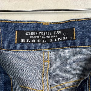 K&ouml;nnte beinhalten: Nahaufnahme einer blauen Jeans mit einem schwarzen Etikett. Auf dem Etikett steht "020608 TEARS OF BLEU CRAFTED IN CALIFORNIA BLACK LINE". Die Jeans hat gelbe N&auml;hte und ein strukturiertes Aussehen.